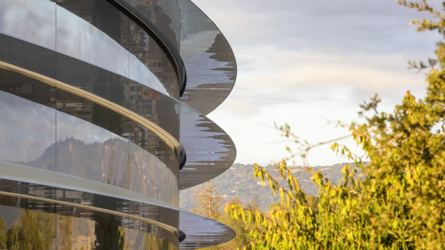 The curved glass exterior of Apple Park in Cupertino, framed by trees.